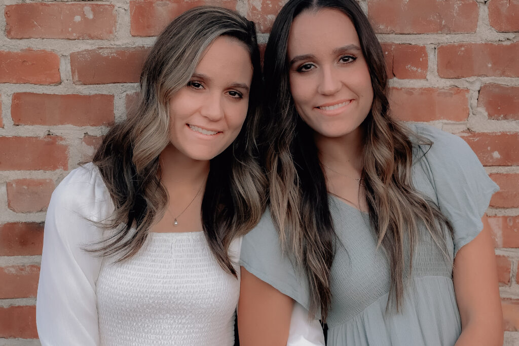 Senior girls wearing a white dress and mint colored dress while leaning up against a brick arms intwined while smiling on for the camera.