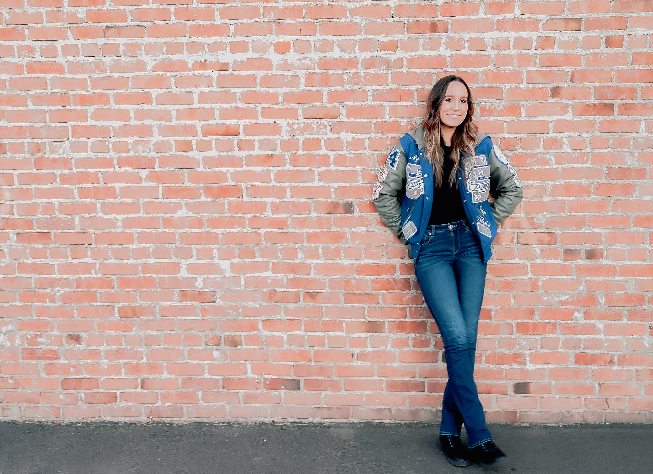 A senior girl wearing her letterman jacket with her hands behind her back on a brick wall while she smiles on.