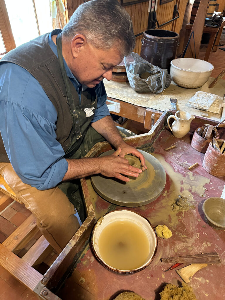A man sitting at a pottery wheel teaching others how to spin clay. 