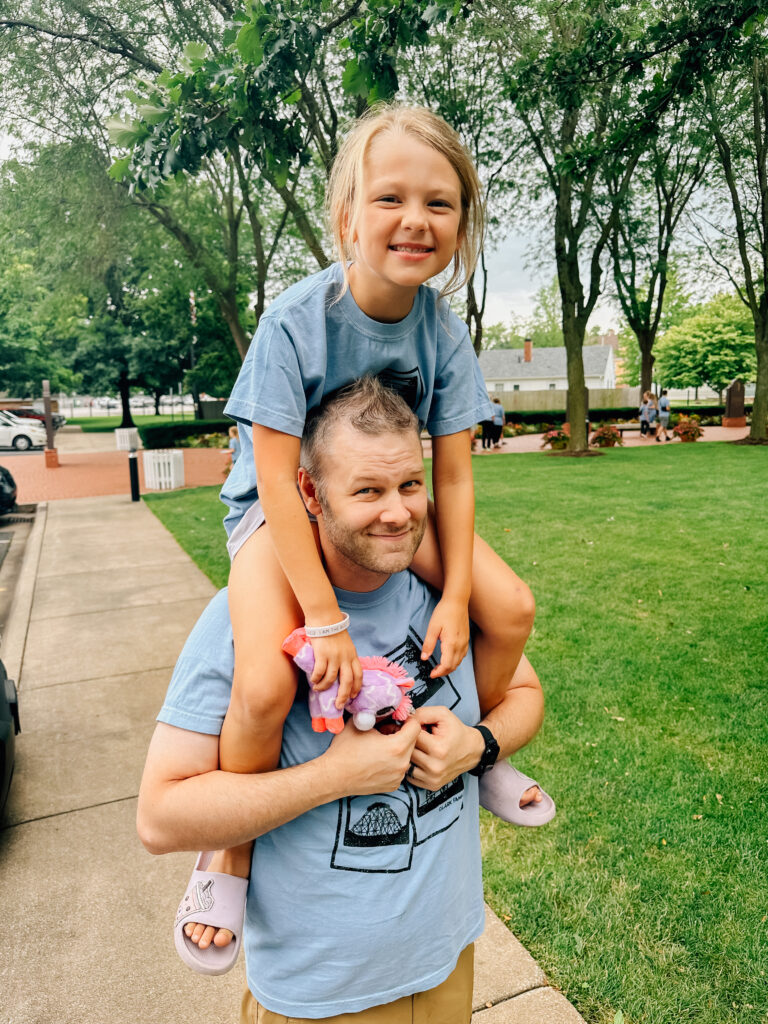 A dad with a little girl on his shoulders while walking on a sidewalk.