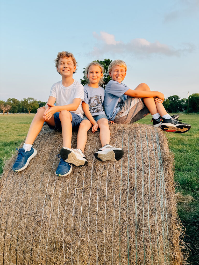 Three kiddos sit excitedly on top of a bale of hay in the middle of a grassy field. 