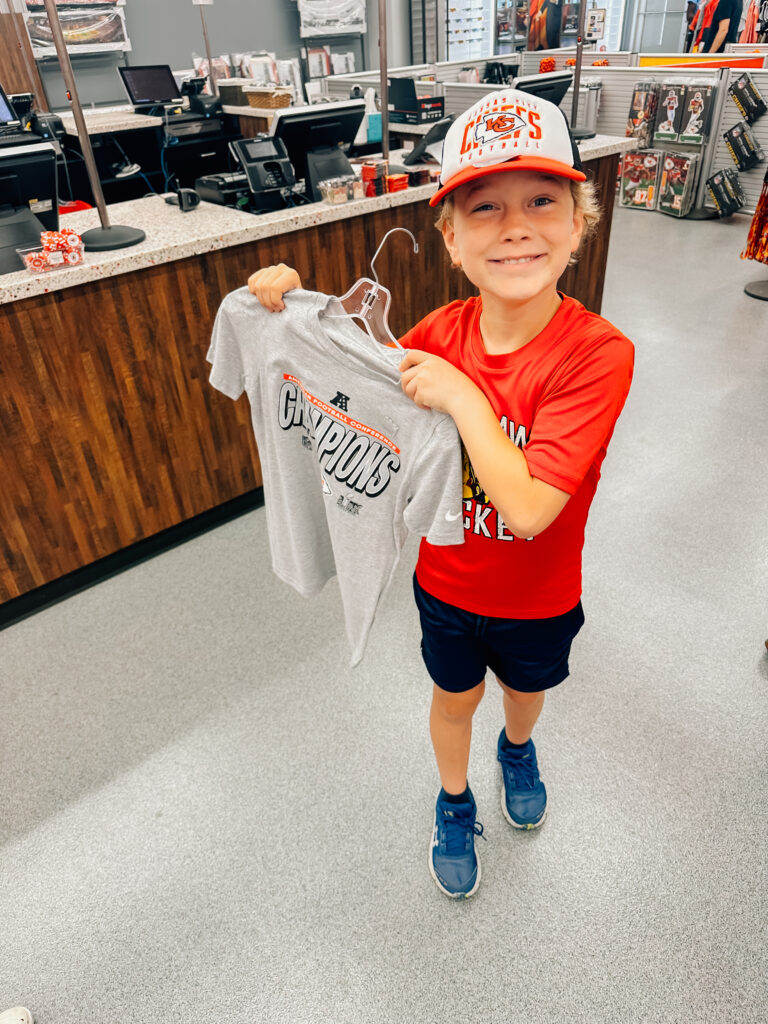 A little boy smiles wide showing off his purchases from the Kansas City Chiefs stadium. A hat on his head and new t-shirt in his hand.