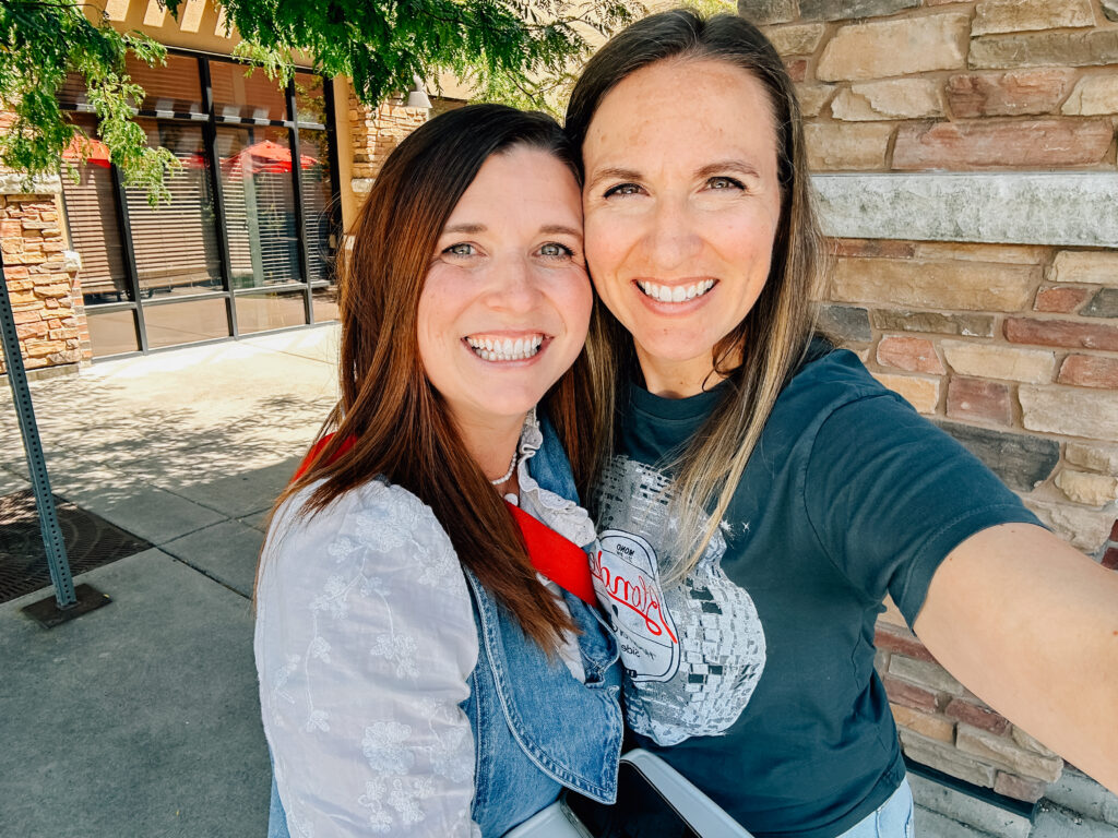 Two women smiling for a selfie while one arm is wrapped around the back of each other. 