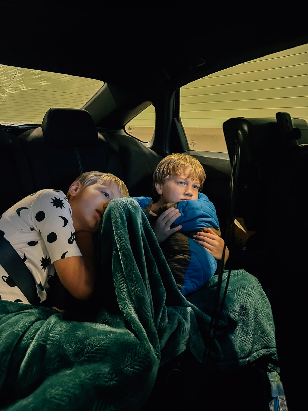 Two boys watch a movie while laying on one another in the back seat of a car while the sunsets behind them. 