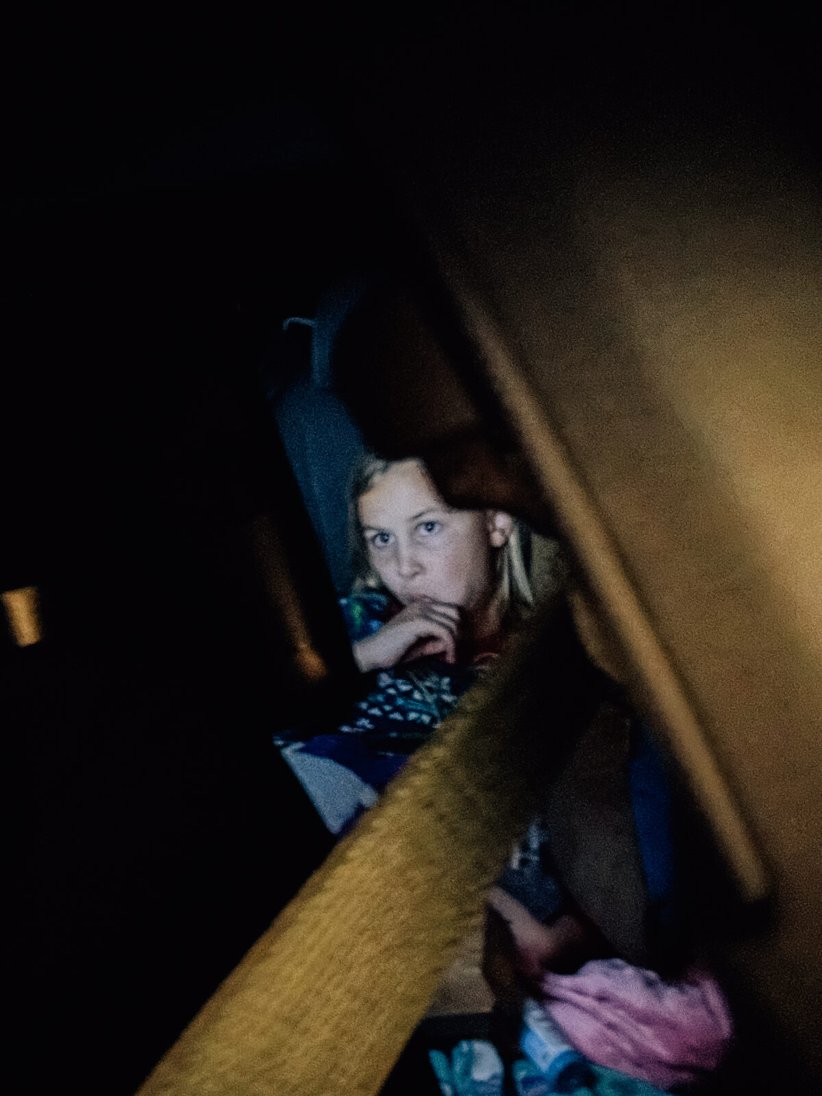 A little girl has her finger in her mouth as she watches a movie in the back seat of the car as the sun goes down. 