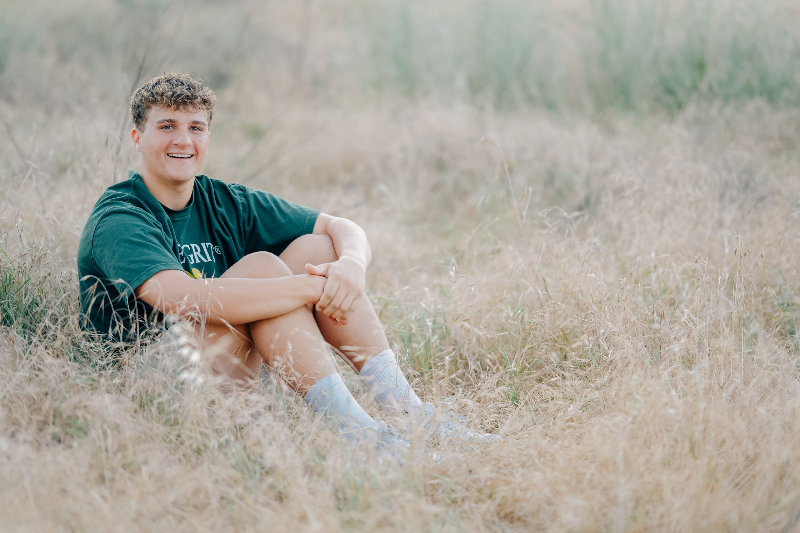 Graduate smiling on while sitting in a field of tall golden grass during a sunset shoot.