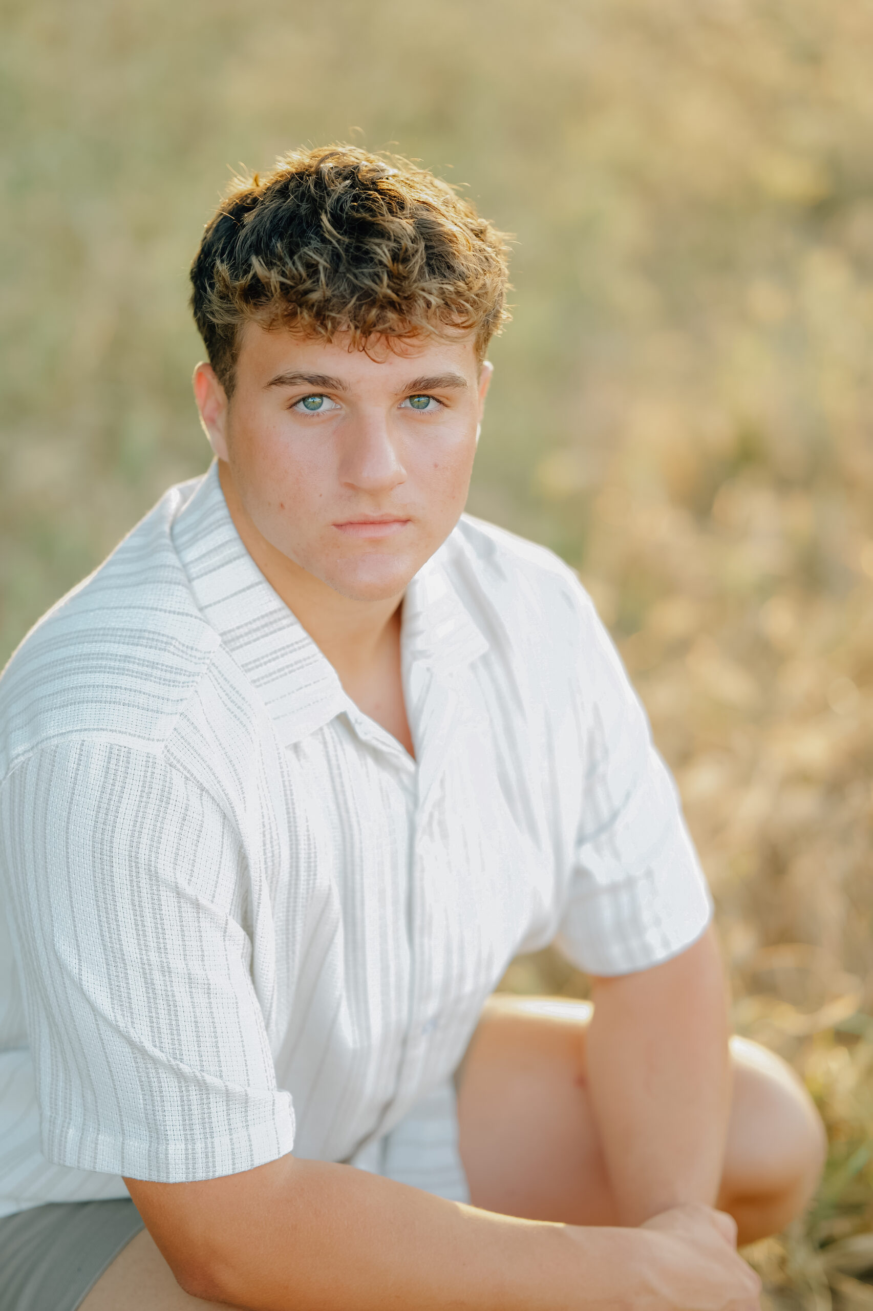 A graduate kneels in tall grass while looking straight on to the camera with a straight face and blue eyes.