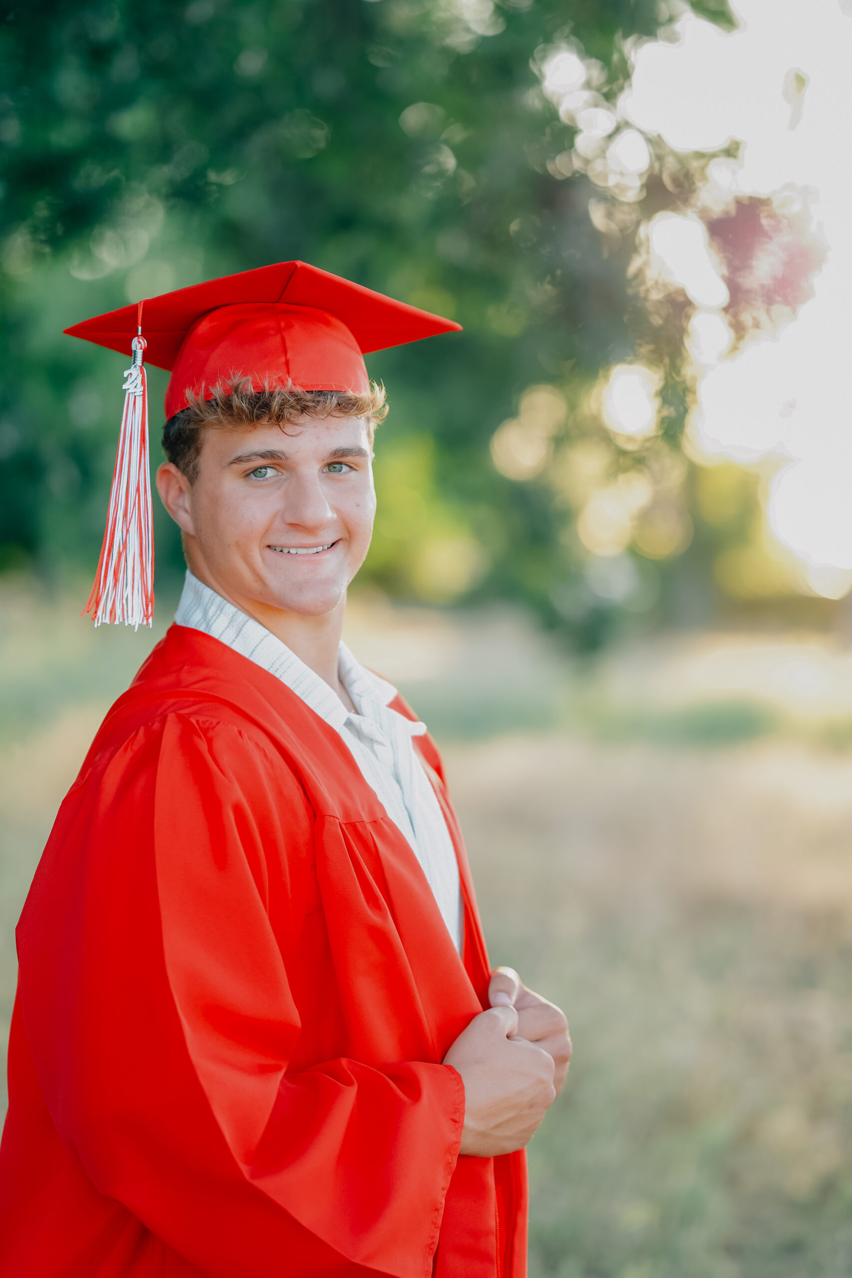 Graduate smiling on while standing holding his robes closed in a field of tall golden grass dressed in his graduation robes during a sunset shoot.