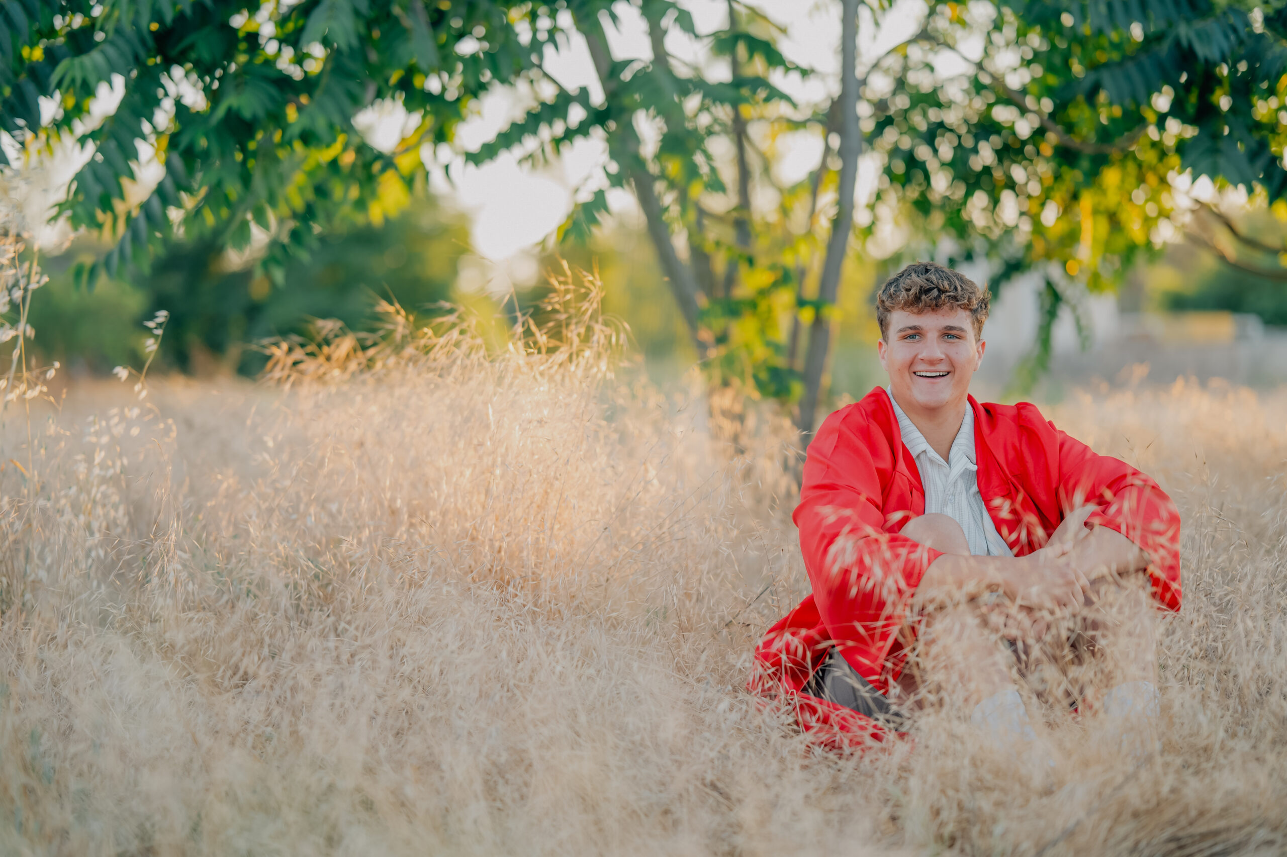 Graduate smiling on while sitting in a field of tall golden grass dressed in his graduation robes during a sunset shoot.