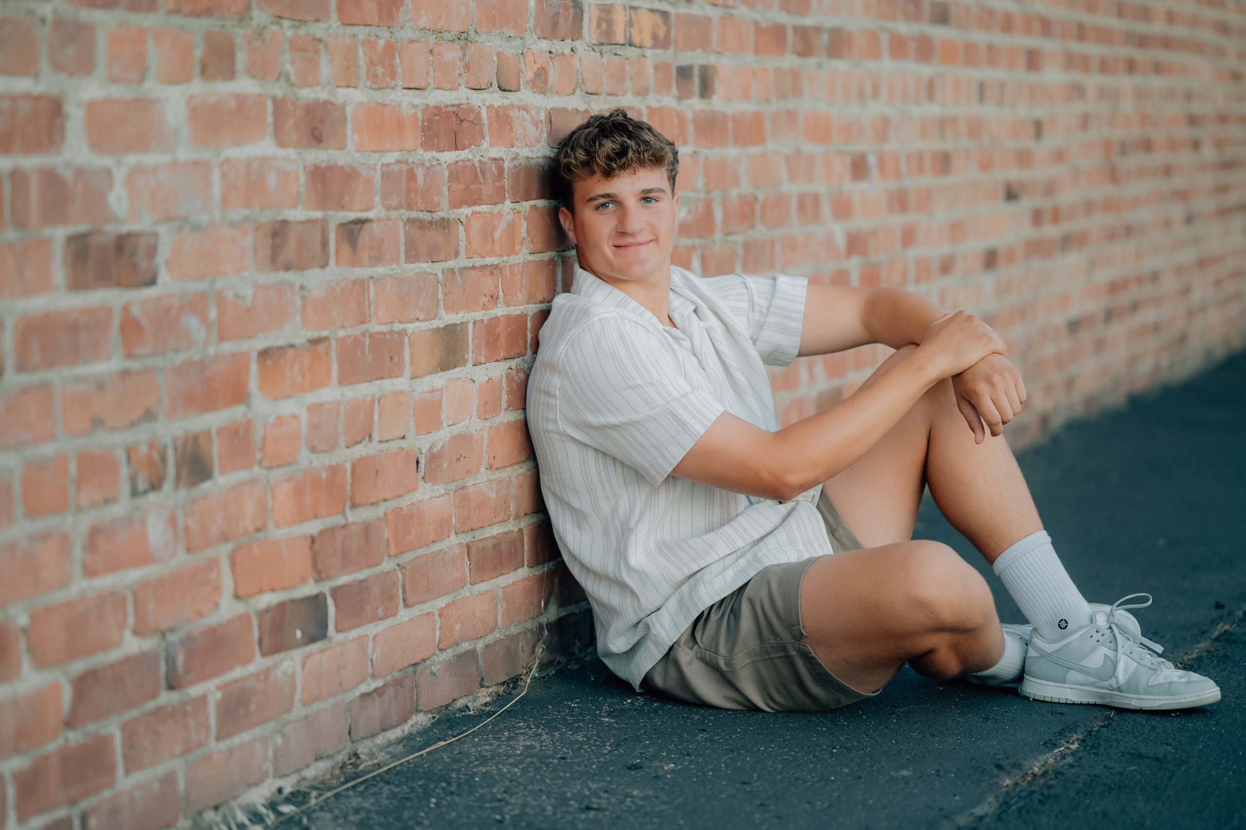 Graduate sits next to a brick wall while looking on at the camera.