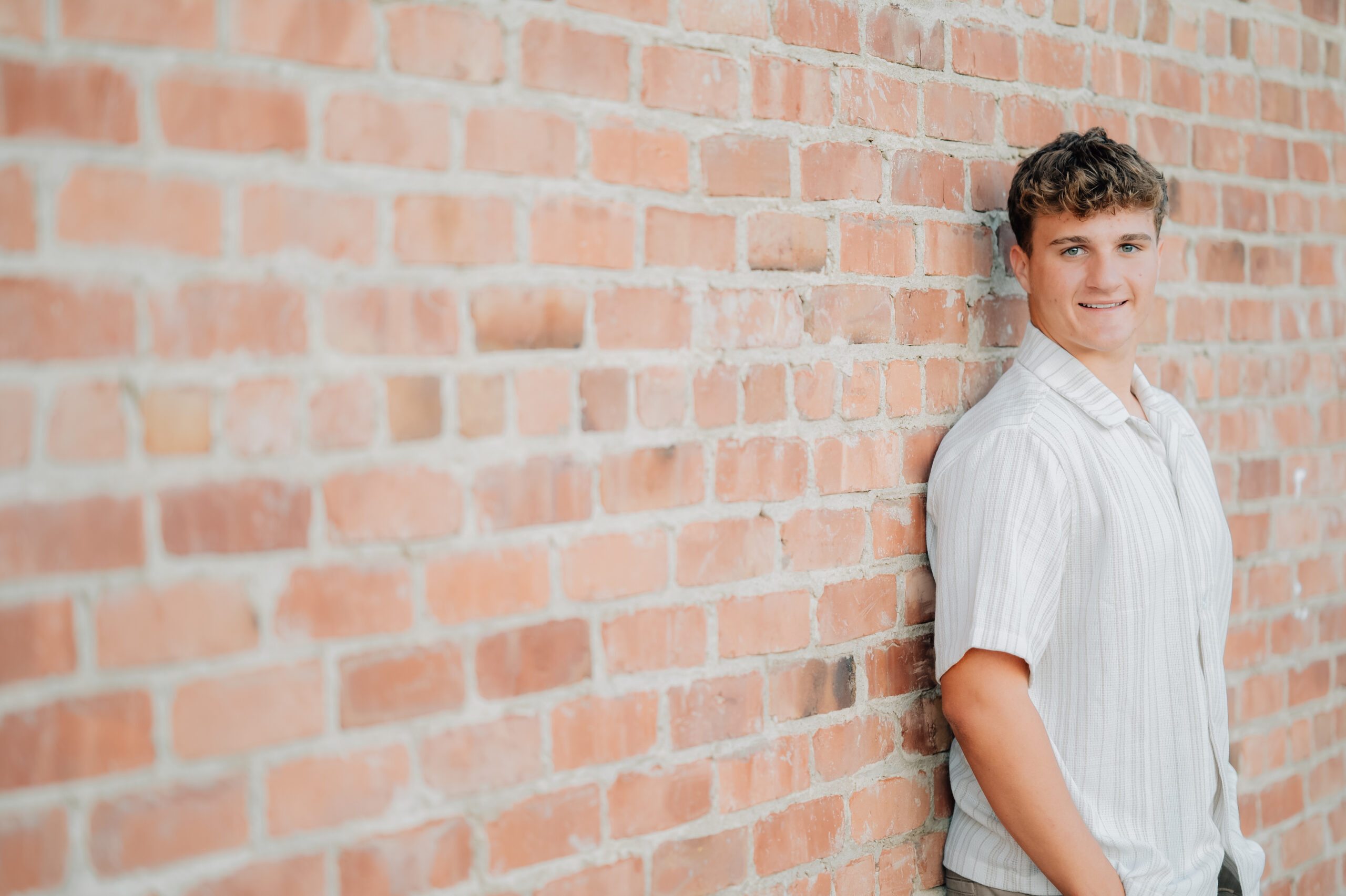 Graduate standing with hands in pockets next to a brick wall while looking on at the camera.