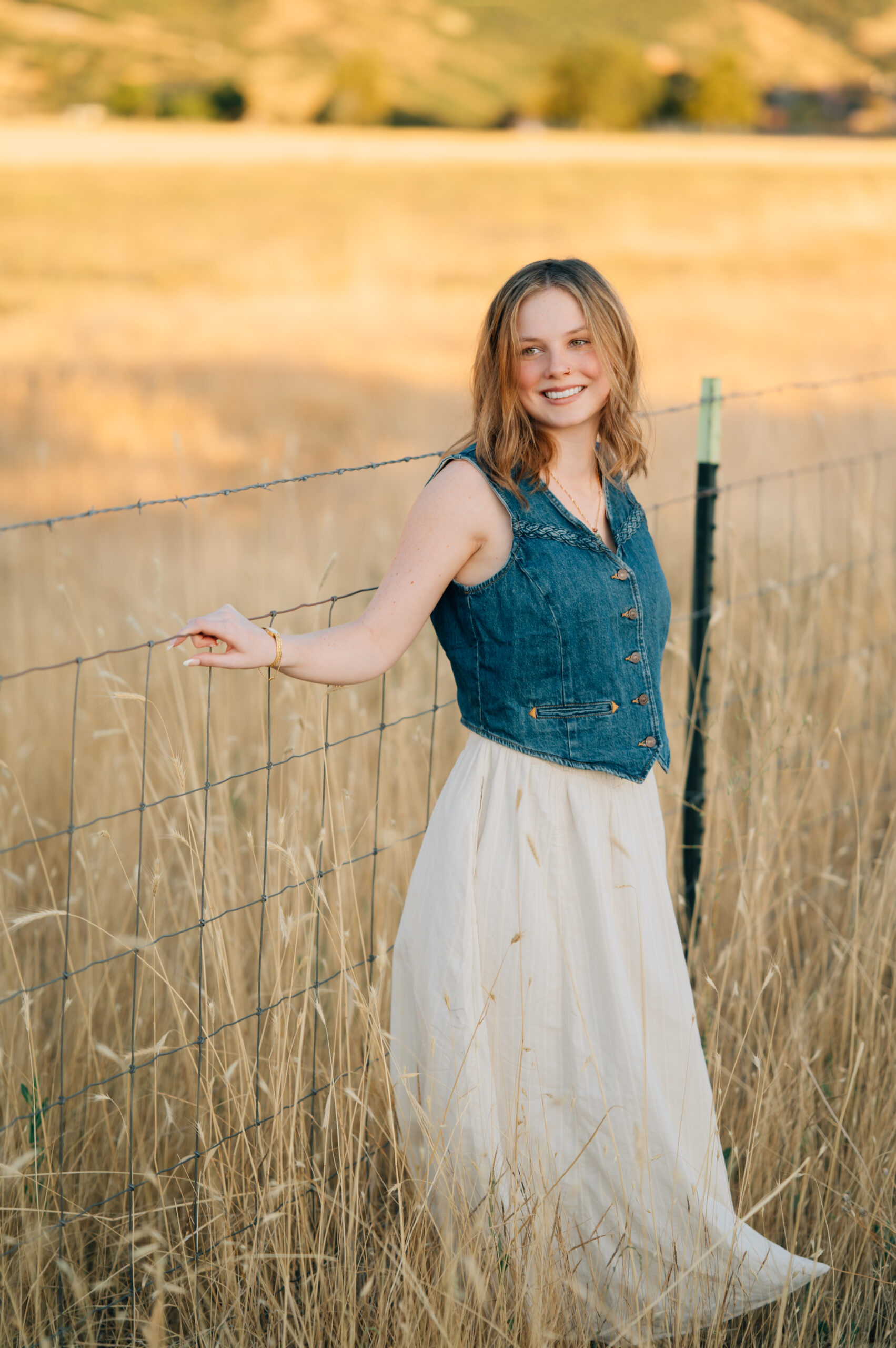 A girl looks to the down and to the side while smiling at a barbed wire fence in tall grass.