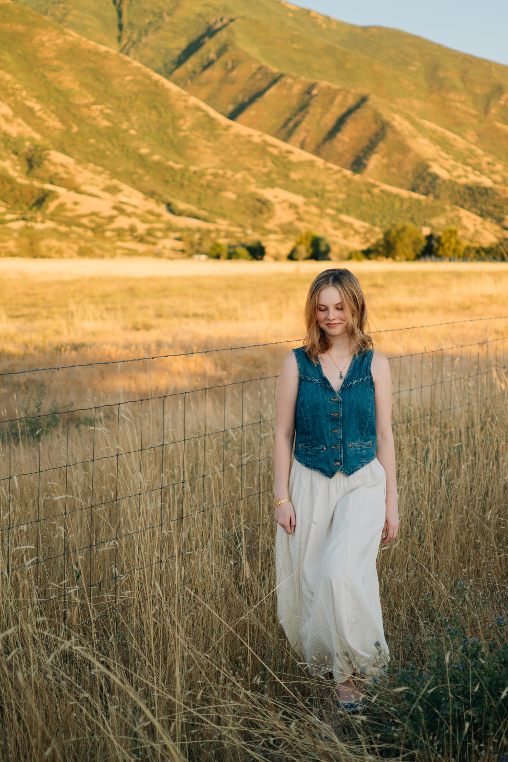 A girl walks along a barbed wire fence through tall grass in the golden light.