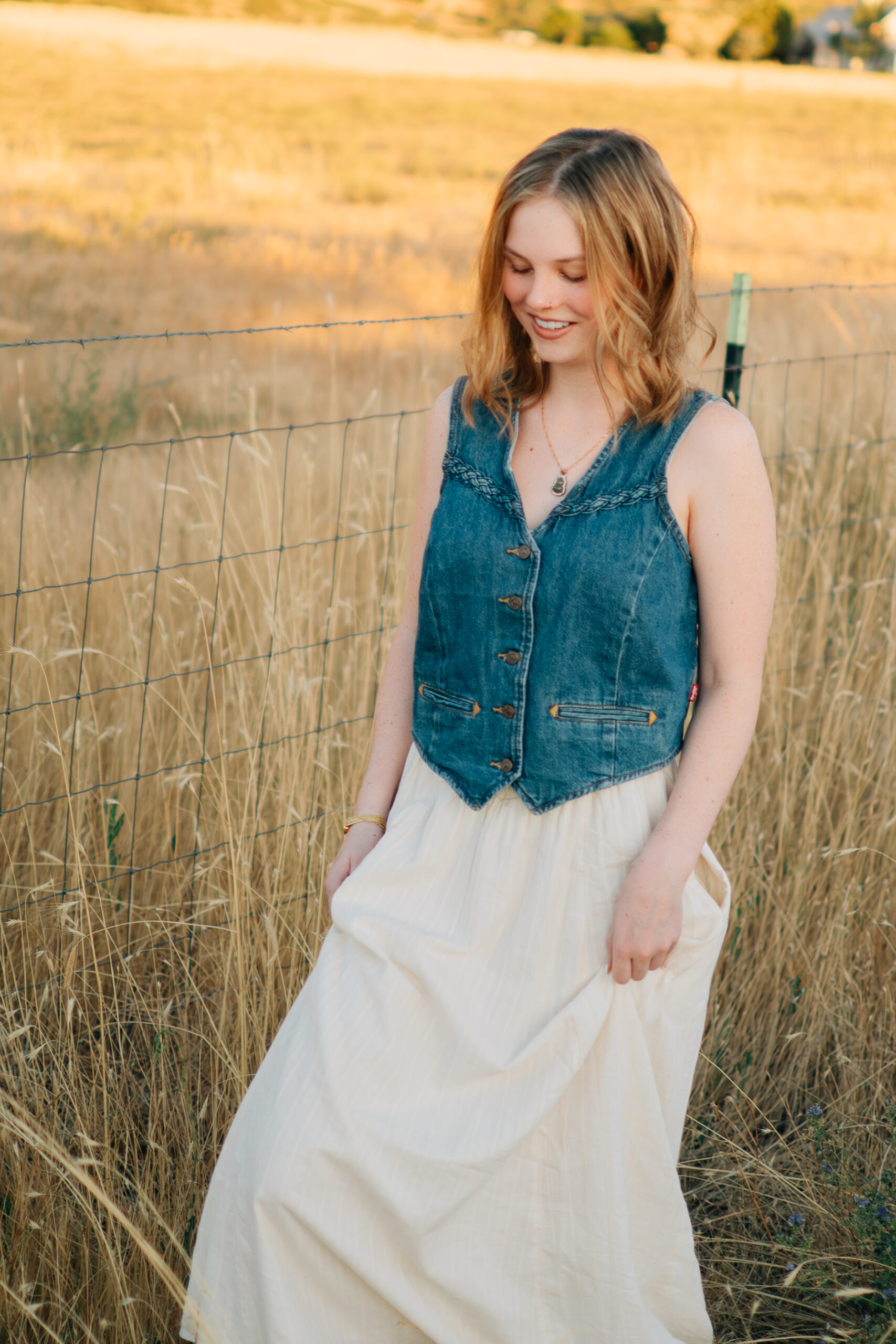 A girl walks along a barbed wire fence through tall grass in the golden light.