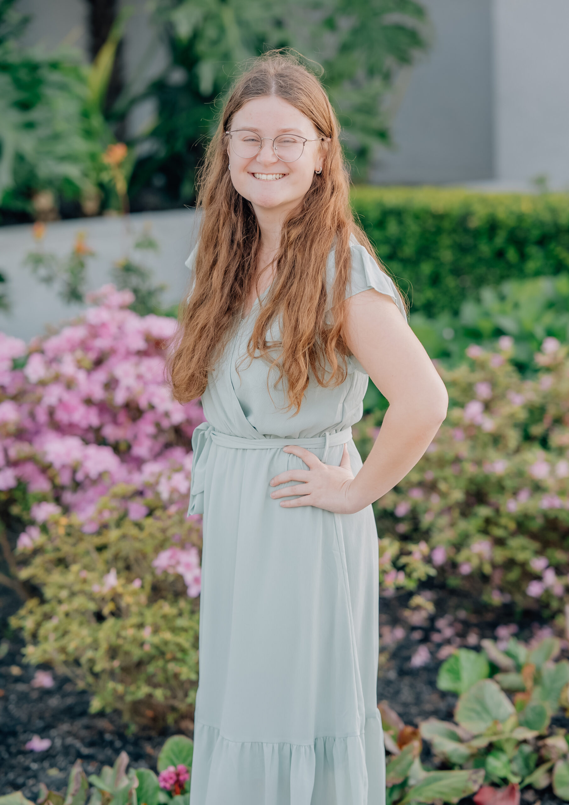 Senior girl in sage green dress smiling in front of pink flowers at the Oakland Temple grounds.