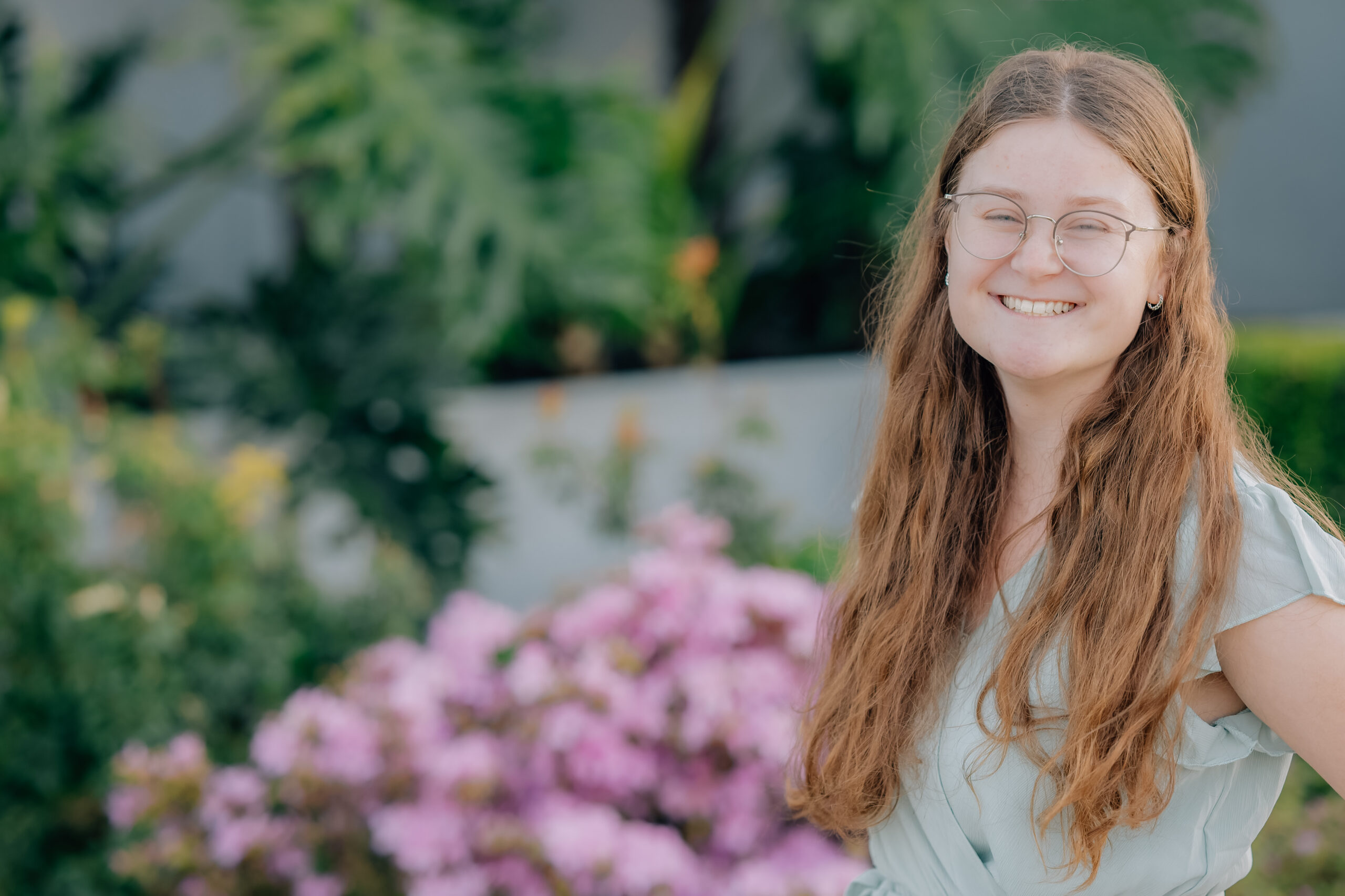 Senior girl in sage green dress smiling in front of pink flowers at the Oakland Temple grounds.