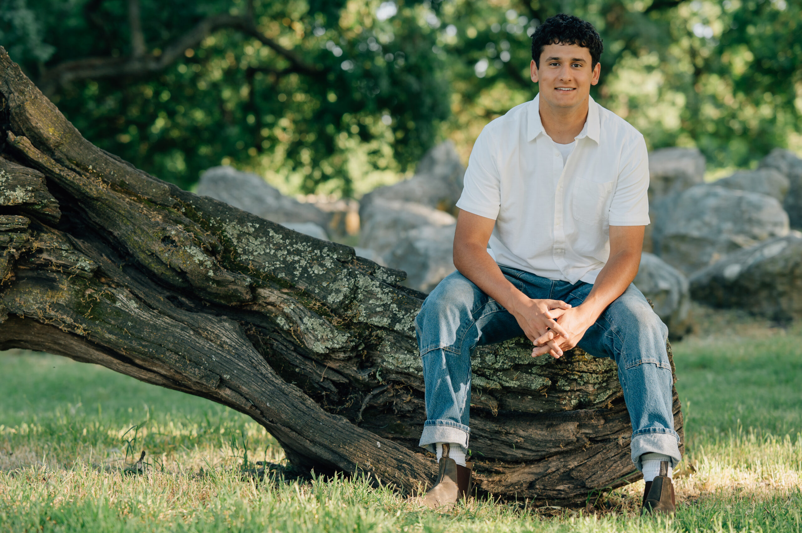 Senior boy at Shumway Oak Grove Park in Stockton sitting on a tree trunk and smiling