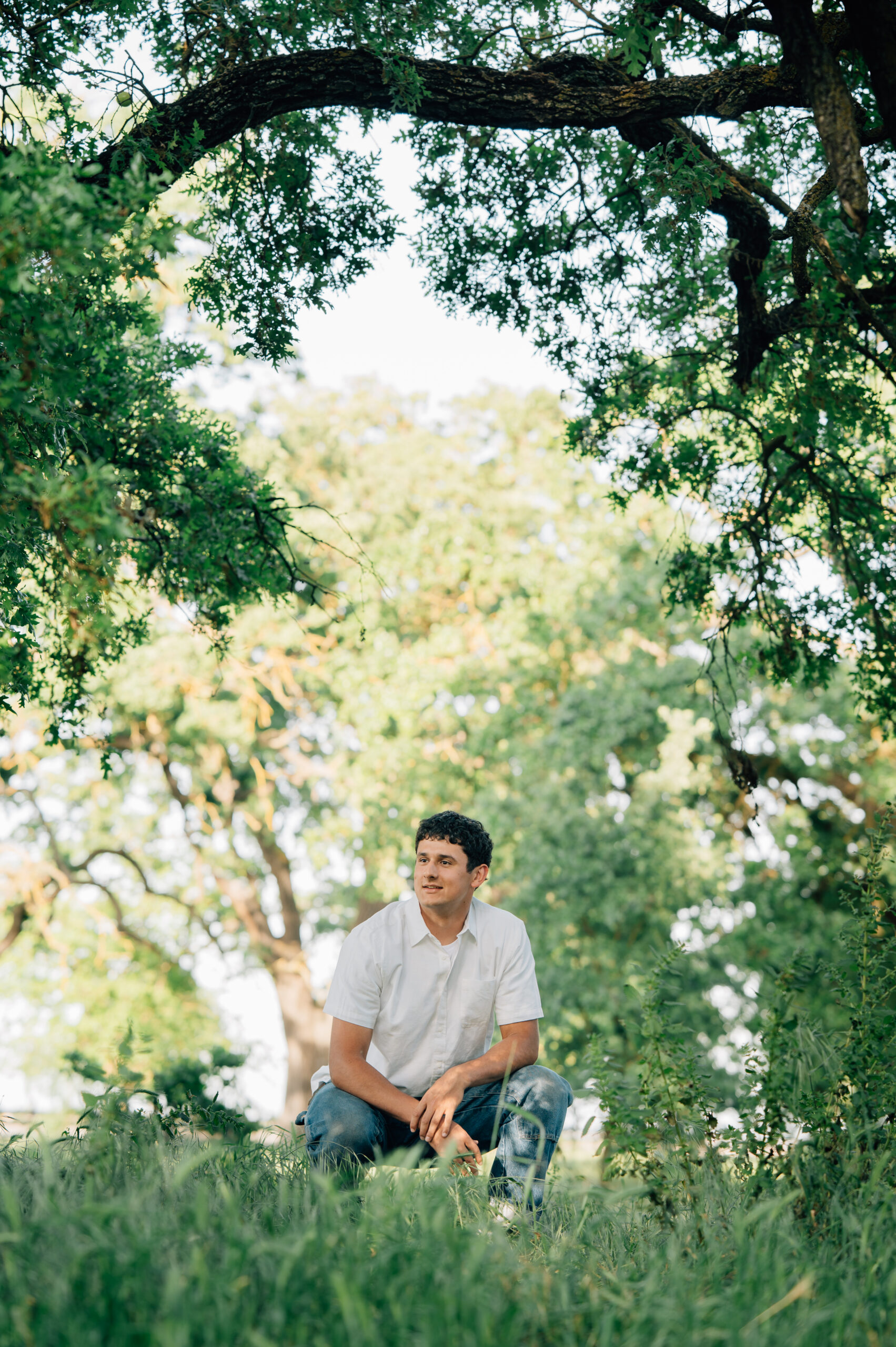 Senior boy kneeling in high grass at Shumway Oak Grove Park in Stockton while looking off to the right.