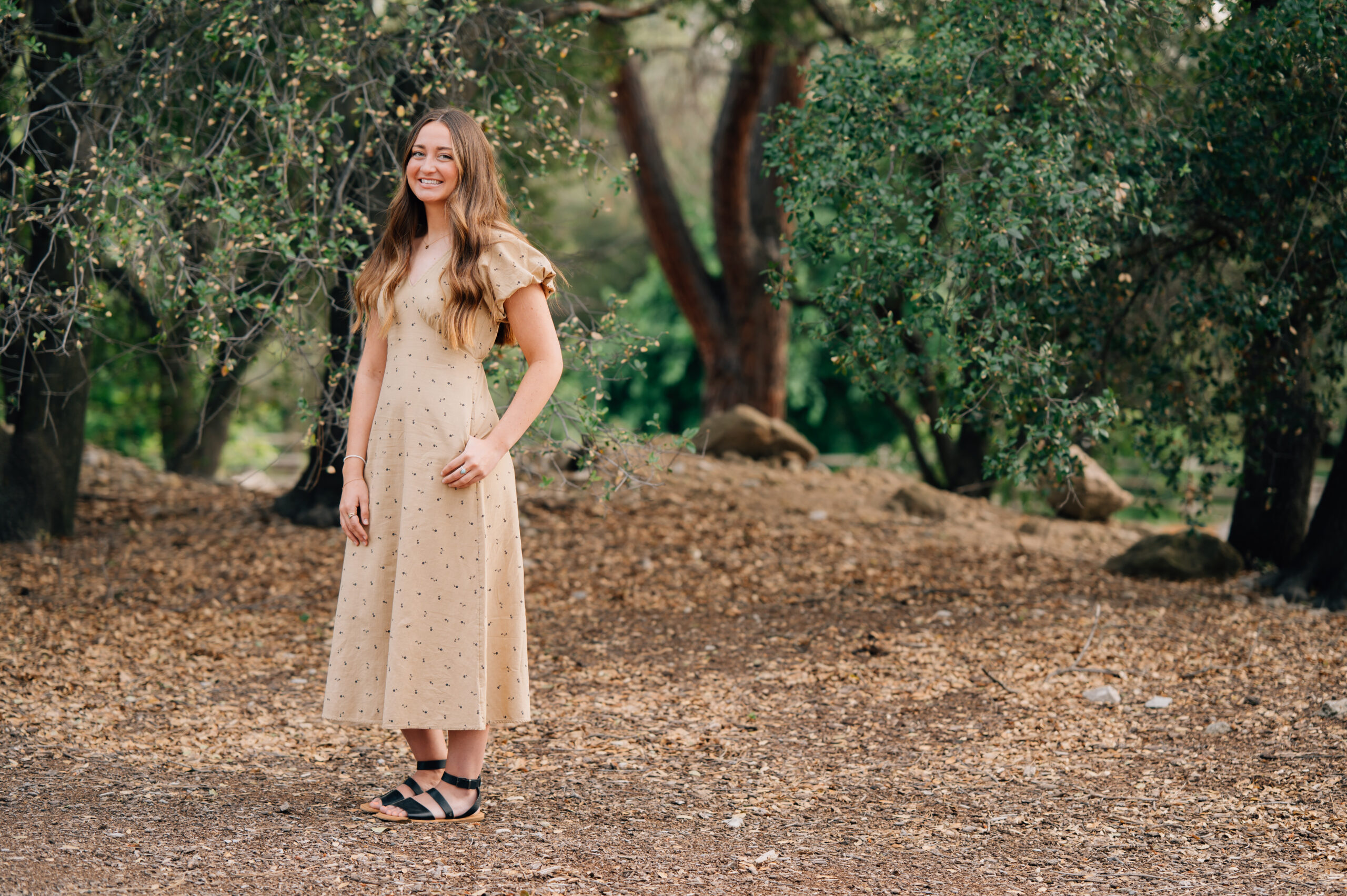 Senior girl posing naturally in tan dress surrounded by trees at Quarry Park