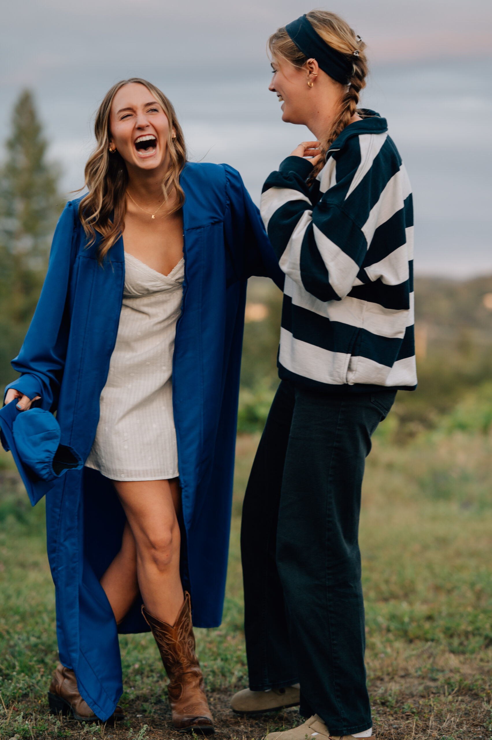High school graduate wearing blue cap and gown laughing with her bestie during outdoor senior portraits in Penryn.