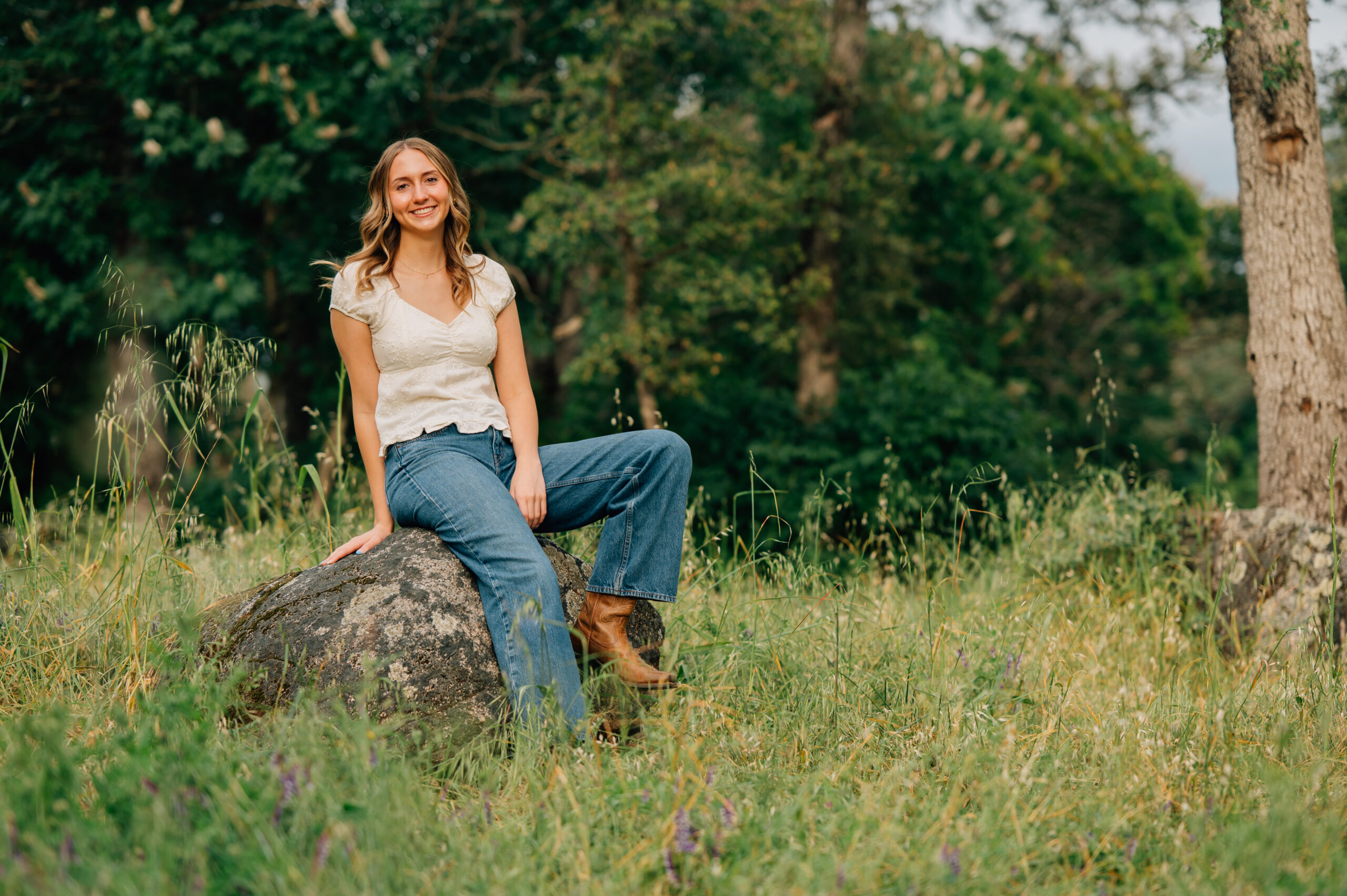 Natural senior portrait of graduate sitting on a boulder in wild grass and smiling during a golden hour session in Penryn.