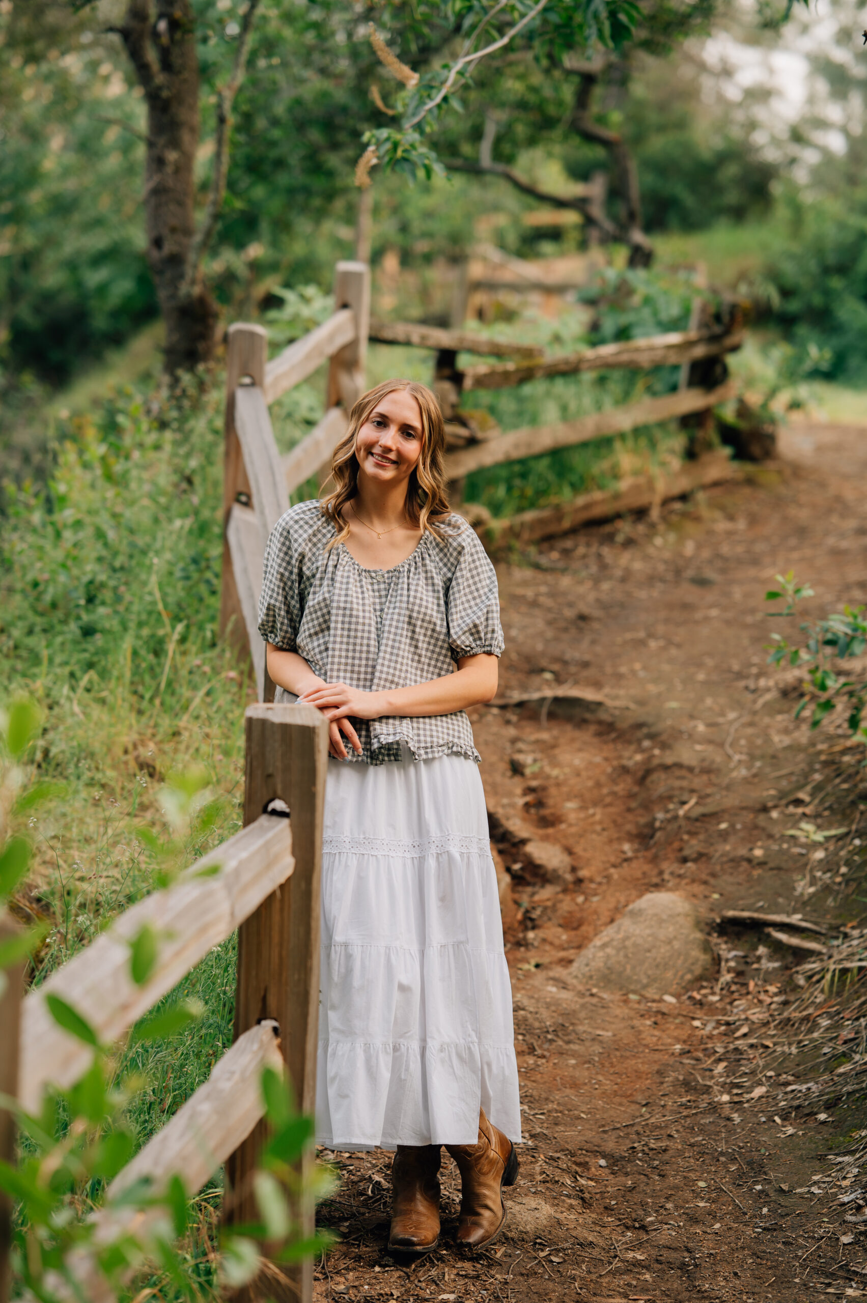 Senior girl in white skirt and cowboy boots standing beside a rustic trail fence at sunset in Northern California.