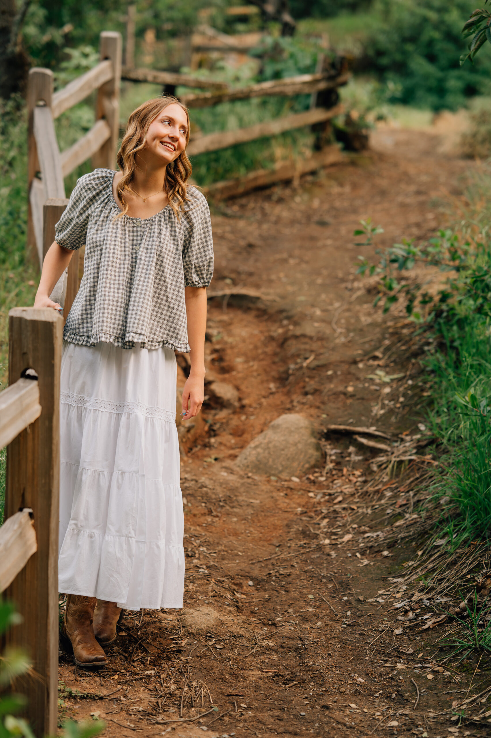 Senior girl in white skirt and cowboy boots standing beside a rustic trail fence at sunset in Northern California.