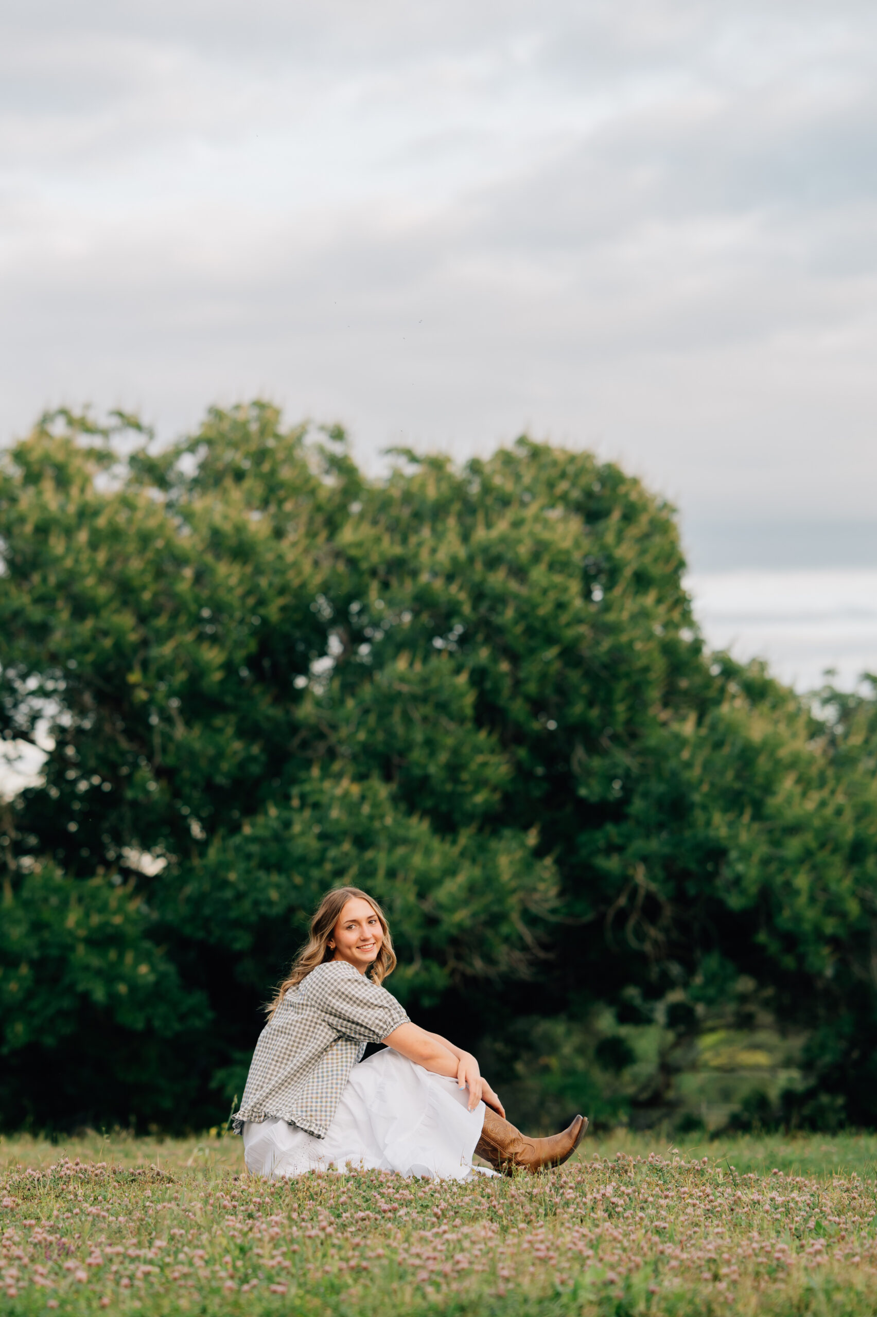 Senior girl with curled hair posing in soft green foliage during an outdoor senior photography session.
