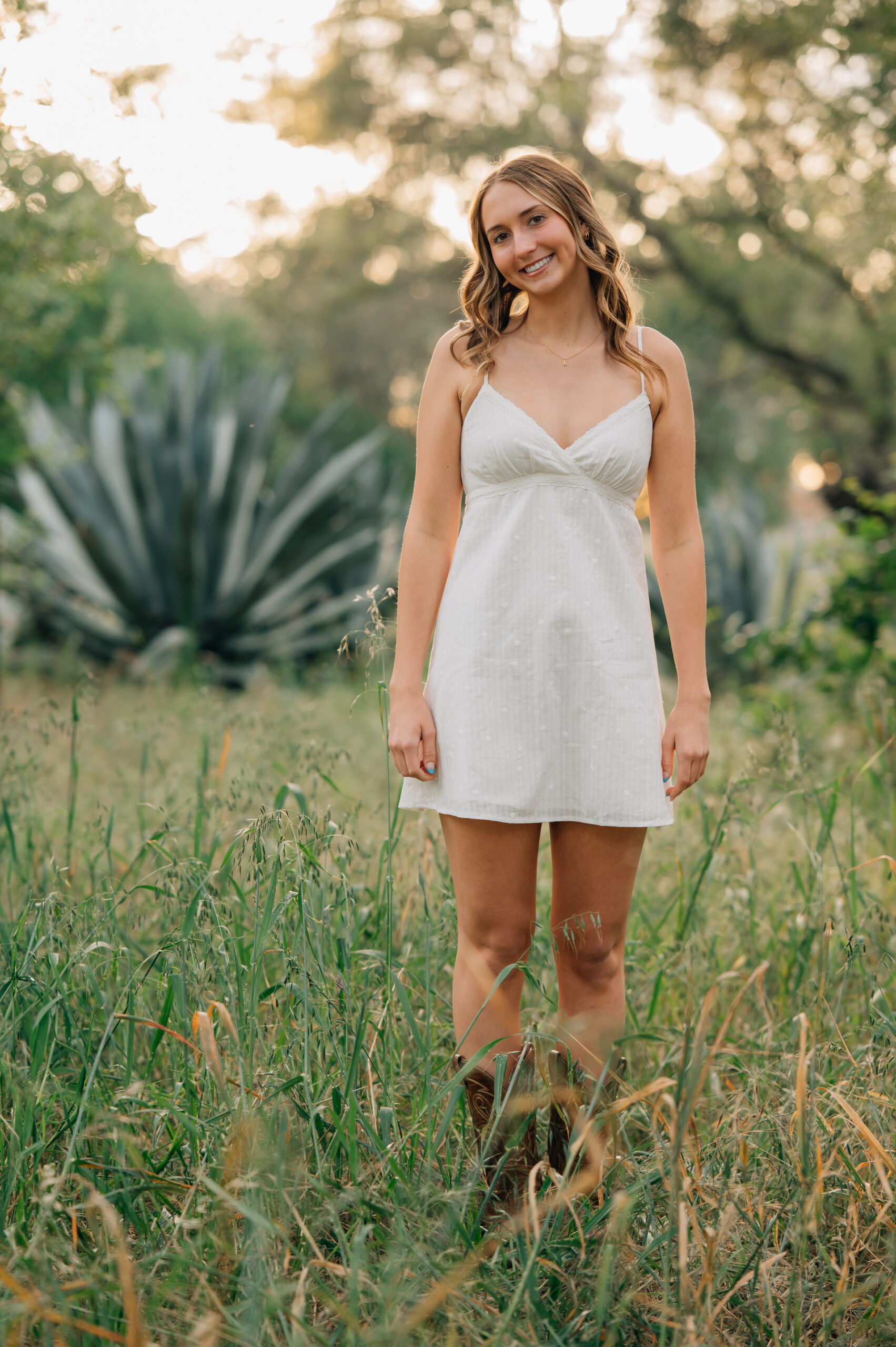 Senior girl in white dress and cowboy boots standing in front of an aloe bush at sunset in Northern California.