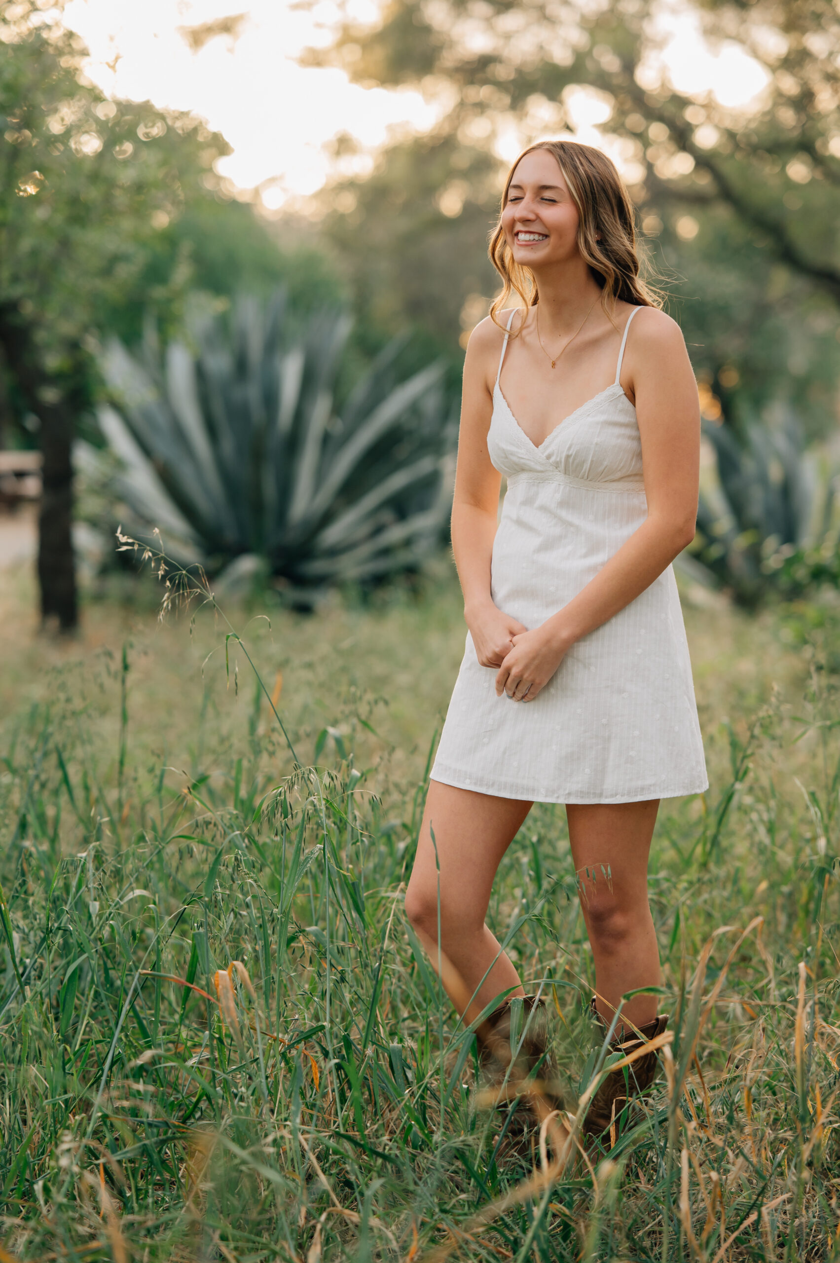 Senior girl in white dress and cowboy boots standing in front of an aloe bush at sunset in Northern California.