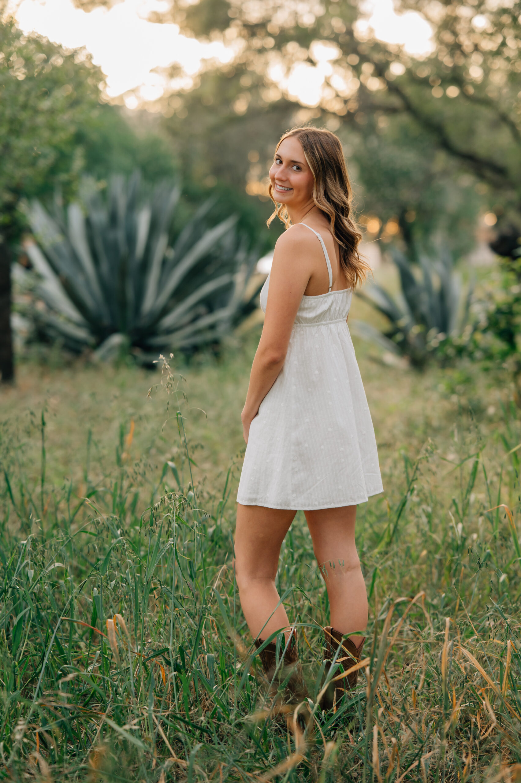 Senior girl in white dress and cowboy boots standing in front of an aloe bush at sunset in Northern California.