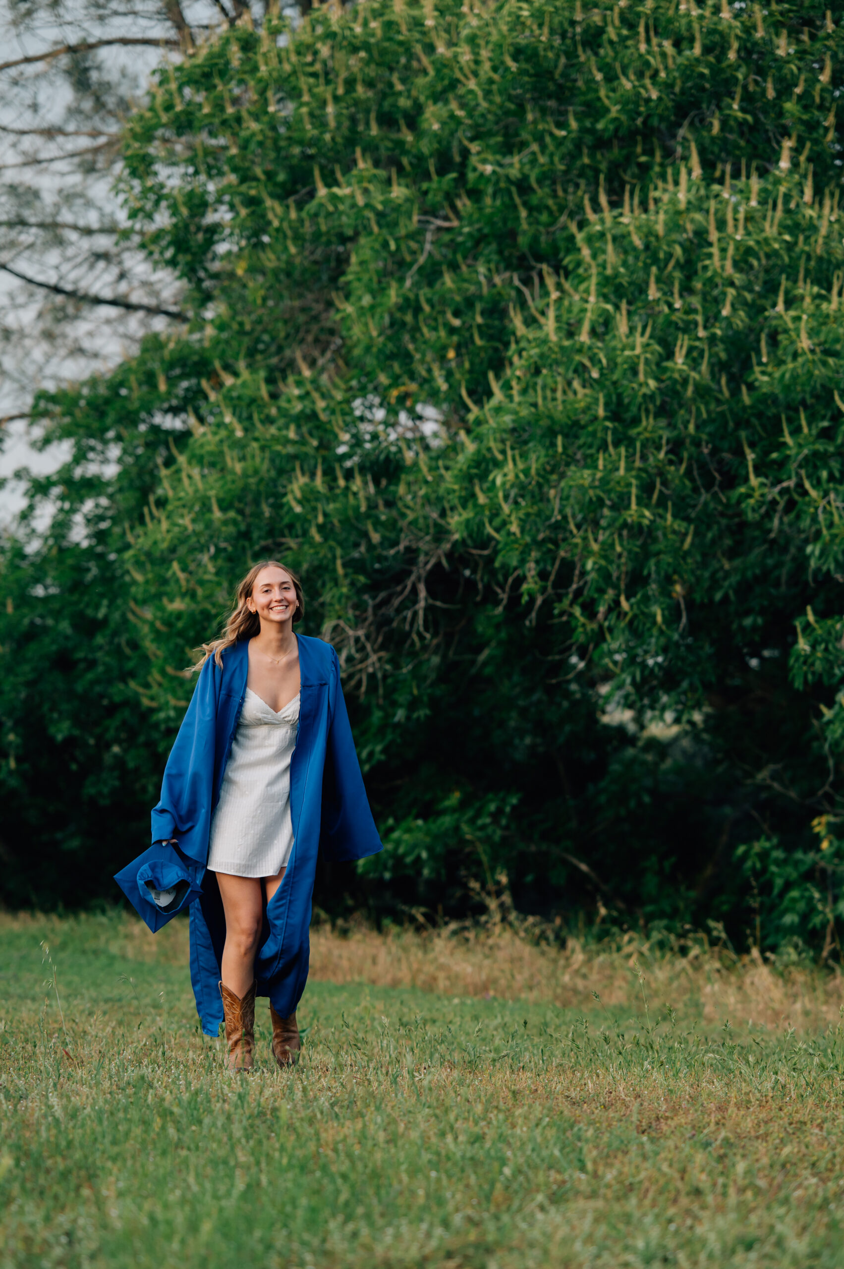 High school graduate wearing blue cap and gown surrounded by greenery and walking during outdoor senior portraits in Penryn.