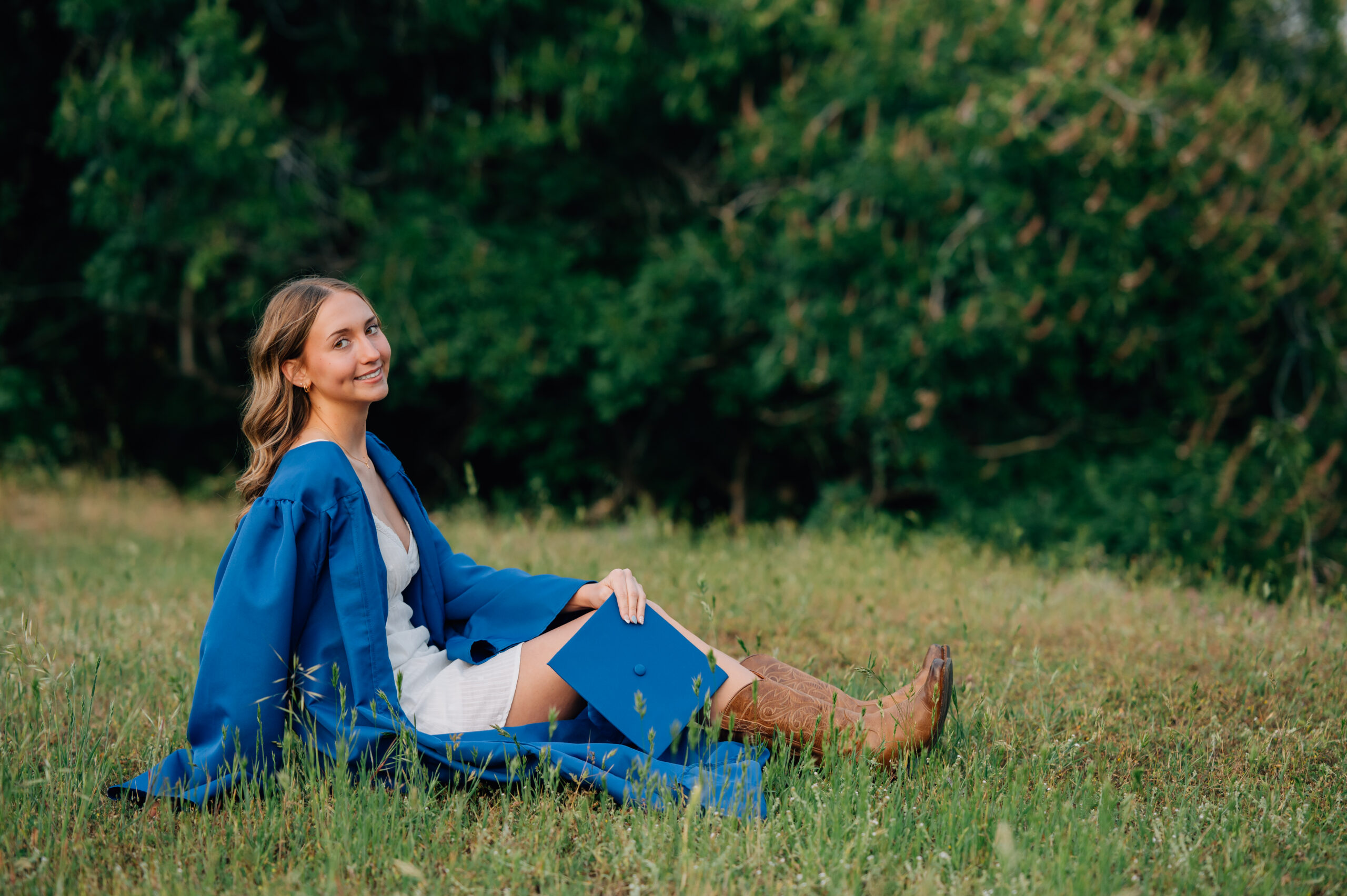 Outdoor graduation portrait of senior girl in royal blue cap and gown standing in front of blooming greenery.