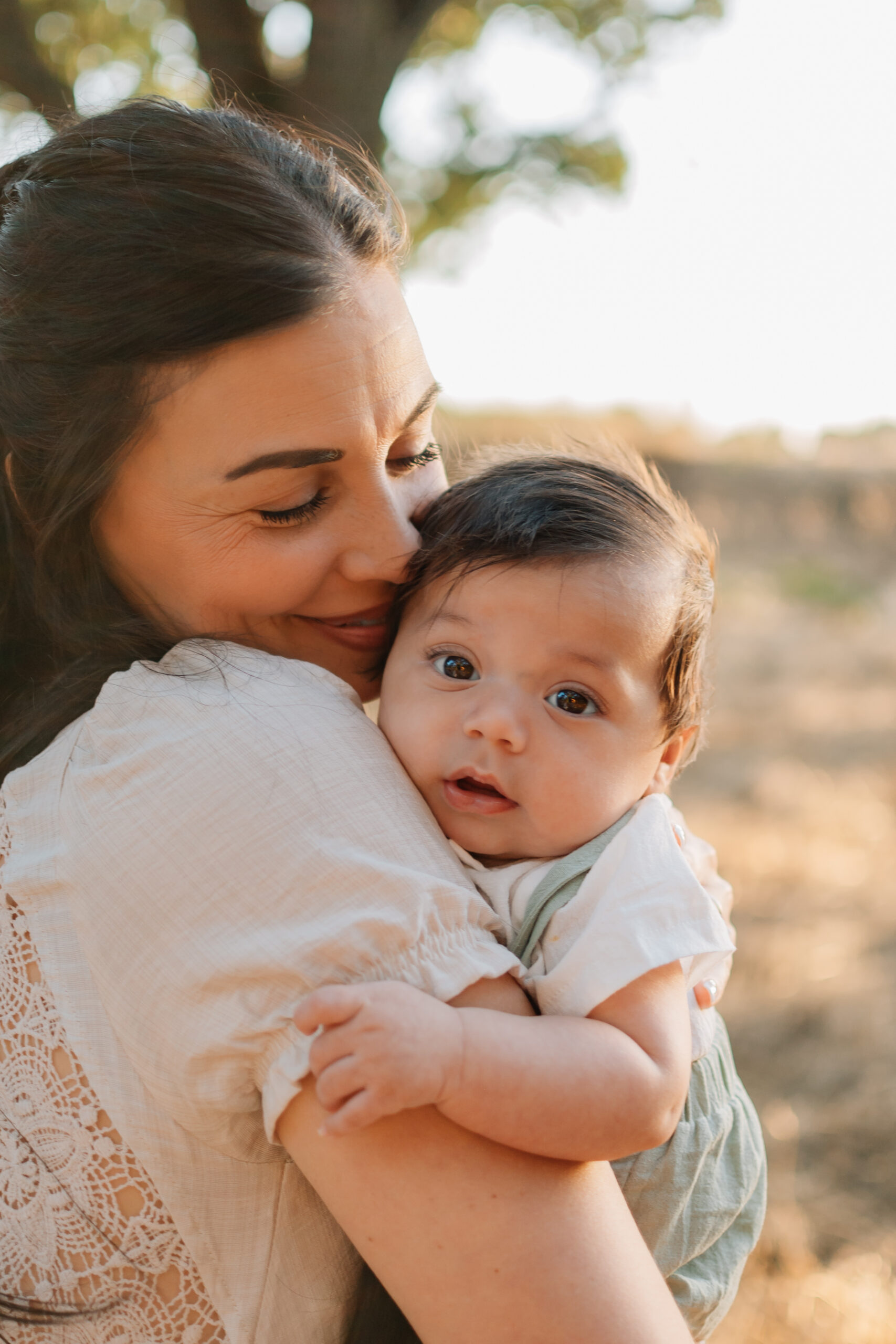 Mother cuddling and kissing her baby in soft golden-hour light at Oak Grove Park.