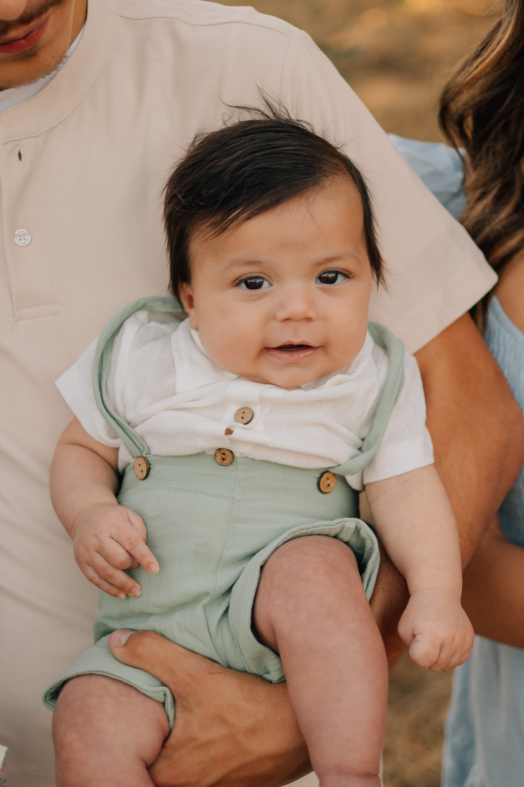 Smiling baby wearing sage green overalls during an outdoor extended family photography session.